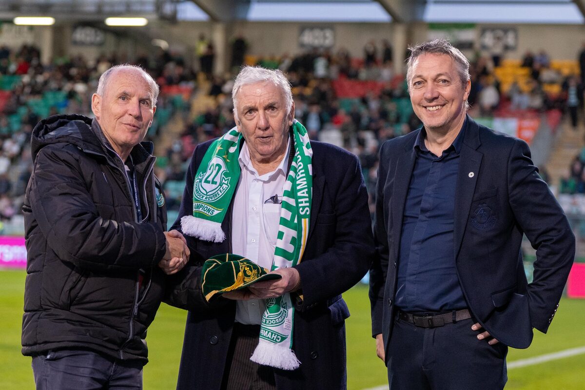 Paddy Mulligan, in a Shamrock Rovers scarf, shaking hands on the pitch at Tallaght Stadium while holding a green and gold Heritage Trust cap, with another man alongside.