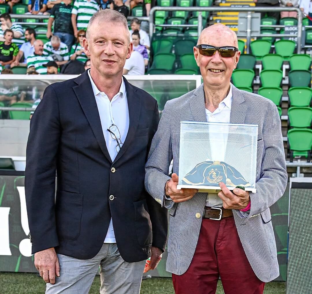 Pat Courtney holding a cased green and gold Heritage Trust cap on the pitch at Tallaght Stadium, with another man alongside him.