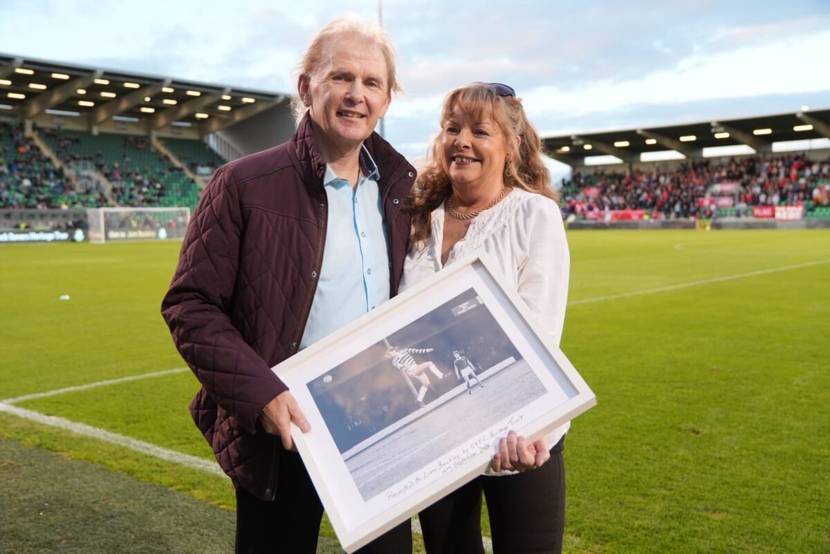 Liam Buckley and a woman holding a framed black and white action photograph of him playing for Shamrock Rovers, on the pitch at Tallaght Stadium.